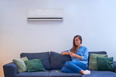 Genç, mutlu bir kadın, klima altında koltukta oturuyor ve sıcaklığı modern evdeki uzaktan kumandayla ayarlıyor.