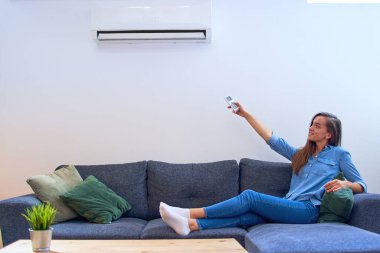Genç, mutlu bir kadın, klima altında koltukta oturuyor ve sıcaklığı modern evdeki uzaktan kumandayla ayarlıyor.