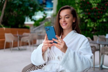 Beyaz bornoz giyen genç ve çekici bir kadın spa merkezinde dinlenirken video izlemek ve internette gezinmek için akıllı telefon kullanıyor.          