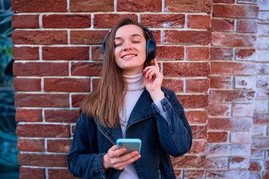 Modern gülümseyen mutlu, sıradan bir genç kadın, açık havada müzik dinlemek için kablosuz kulaklık ve akıllı telefon kullanıyor.