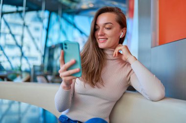 Genç, modern, neşeli, kablosuz kulaklık takan, video izlemek için akıllı telefon kullanan ve kafede oturup dinlenirken internetten haber okuyan bir kadın. 