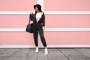 Tarz sahibi, genç, sevimli, siyah tulumlu, panama şapkalı, kısa beyaz bluzlu, spor çantası tutan ve parlak pembe bir duvarın karşısında duran bir kadın.