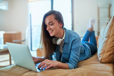 Kablosuz kulaklıklı, gündelik akıllı, modern gülümseyen kız video içeriğini izlemek için laptop kullanıyor, sosyal medyayı kontrol ediyor ve evdeki kanepede dinlenirken internetten alışveriş yapıyor.