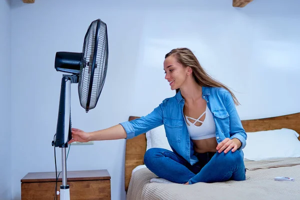Smiling young cute woman sitting on bed in the bedroom in sunny hot summer day and enjoying of fresh air in the front of electric working fan
