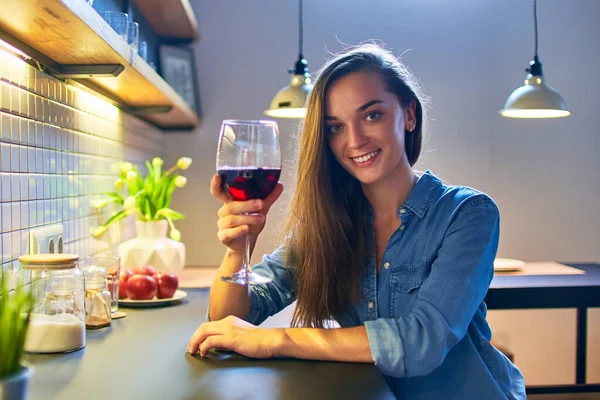 在家里阁楼风格的厨房里 漂亮可爱的年轻而随意的笑容满面的饮酒女士举着红酒坐在餐桌旁的画像 — 图库照片