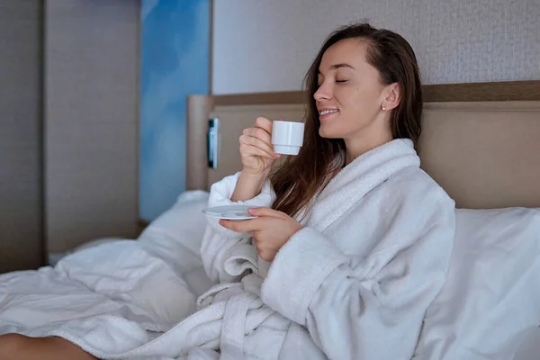 バスローブを着た怠惰な女性がベッドの上に寝そべって ホテルの部屋で居心地の良い快適なベッドルームでリラックスしている間 香り豊かなコーヒーカップで朝の朝食を楽しんでいます 簡単な生活 新しい一日の始まり — ストック写真