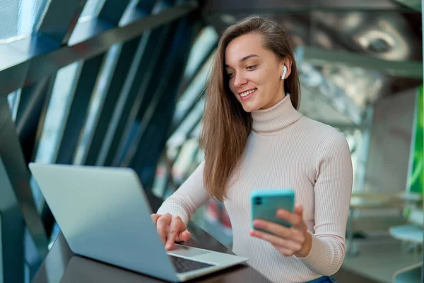 若い現代のカジュアルな幸せなスマート女性が座ってカフェで休んでいる間にビデオや遠隔作業をオンラインで見るためにノートパソコンや携帯電話を使用して無線ヘッドフォンを身に着けている — ストック写真