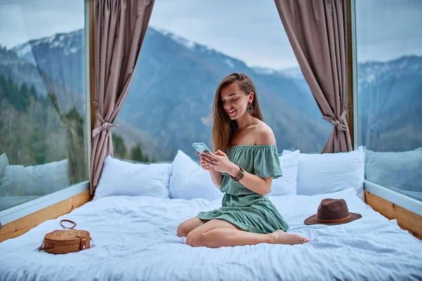 若い魅力的な笑顔美しいBohoシックなブルネットの女性旅行者裸の肩エメラルドドレスを身に着けている山の景色とオンライン通信のための電話を使用してホテルの部屋にベッドに座って — ストック写真