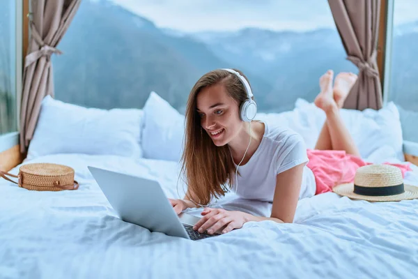 自由女旅行者躺在床上 使用耳机和笔记本电脑进行远程在线工作 通信和观看视频会议 — 图库照片