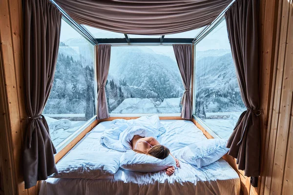 穏やかな穏やかな女性は 雪の多い冬の山々の景色を望む部屋で柔らかい白い居心地の良い快適な暖かいベッドで寝ています — ストック写真