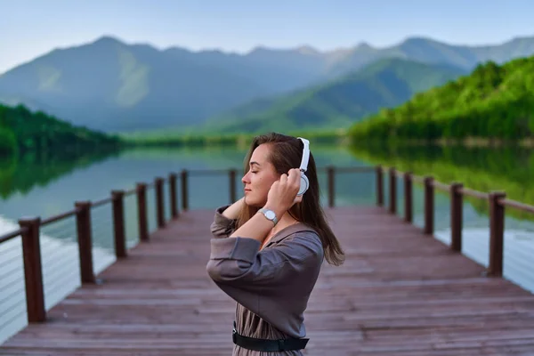 湖と山の景色を望む桟橋に一人で立っている無線ヘッドフォンで穏やかな音楽を聞き 穏やかな静かな静かな静かな雰囲気を楽しむ女の子の旅行者 — ストック写真