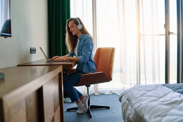 stock image Millennial happy smiling caucasian girl freelancer with wireless headphones sit at desk in hotel room and remote working using laptop. Take online course or training at home