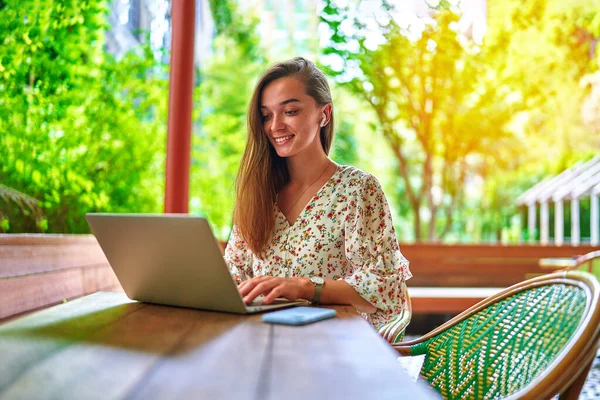 幸せな陽気な笑顔美しいかわいい楽しい若い千年紀のフリーランスの女の子リモートワークと緑のカフェでオンラインブラウジングのためのラップトップを使用して無線ヘッドフォンを身に着けています — ストック写真