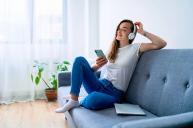 Genç mutlu, mutlu, mutlu, bin yıllık müzik aşığı bir kanepede oturuyor ve evde çevrimiçi müzik dinlemekten zevk almak için kablosuz kulaklık kullanıyor.