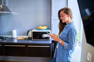 Genç, çekici, gülümseyen, mutlu, sevimli bin yıllık kız pencerenin önünde duruyor ve telefonla internette sohbet ediyor, internette sörf yapıyor, internette geziniyor ve sosyal ağları kullanıyor. 