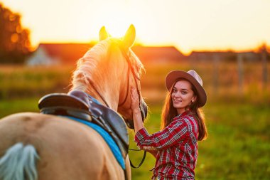 Genç, sevimli, mutlu, mutlu, gülümseyen bir kadın gün batımında çayırda güzel, sarışın palomino atını okşuyor.