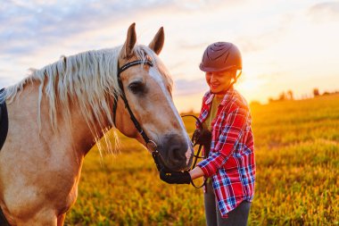 Genç, mutlu, neşeli, mutlu, güler yüzlü, kasklı, sarışın palomino atı okşayan, altın saatinde gün batımında çayırda gülümseyen bir kadın sürücü. 
