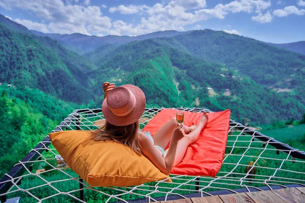 緑の大きな山を背景に無料の幸せな瞬間の生活を楽しんでいる間 若い女の子の旅行者はワインのガラスでリラックスしてハンモックに横たわっている帽子をかぶって — ストック写真