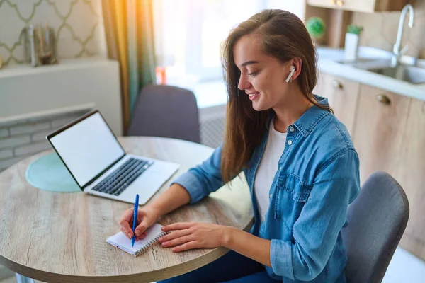 幸せな若いスマート千年紀の女性はノートパッドにペンでノートを取り ワークスペースでオンライン教育のための開いて白い空白のディスプレイとラップトップを使用してノートを取ります ウェブサイトの広告のためのコンピュータのモックアップ — ストック写真
