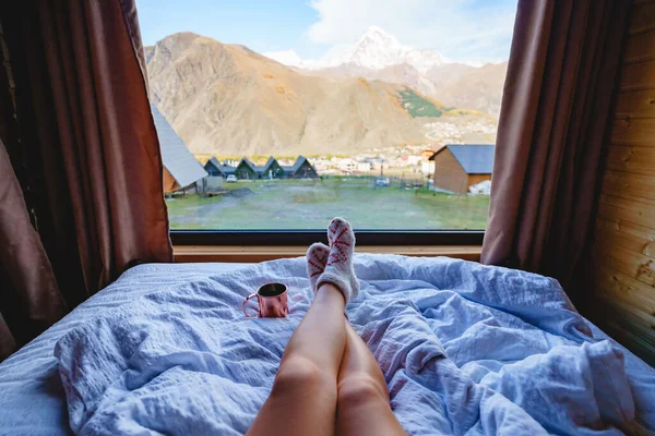 暖かいニットの靴下の長い女性の足は大きな窓と山の景色を望むベッドの上にあります — ストック写真