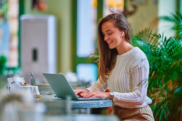 かわいい若い幸せな笑顔スマート現代のコピーライターのフリーランスの女の子は カフェでリモートでオンラインで作業中にノートパソコンのキーボードを入力無線ヘッドフォンを身に着けて — ストック写真