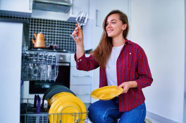 Genç, mutlu, mutlu, gülümseyen bir ev hanımı modern bulaşık makinesini beyaz ev mutfağında bulaşık yıkamak ve bardak yıkamak için kullanıyor. 