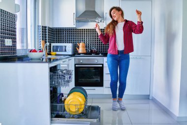 Genç, mutlu, mutlu, gülümseyen, dans eden bir ev hanımı modern bulaşık makinesini beyaz ev mutfağında bulaşık yıkamak ve bardak yıkamak için kullanıyor. Temizlik işlerinin tadını çıkar. 