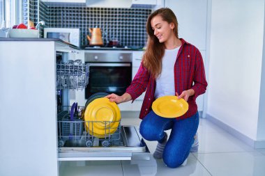 Genç, mutlu, mutlu, gülümseyen bir ev hanımı modern bulaşık makinesini beyaz ev mutfağında bulaşık yıkamak ve bardak yıkamak için kullanıyor. 