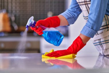 Önlük ve kırmızı koruyucu eldiven giyen ev hanımı toz masasını sprey ve bez kullanarak temizler ve dezenfekte eder. 