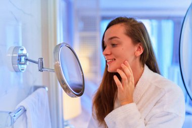 Young smiling girl looks at herself in a small round wall mirror after a shower 