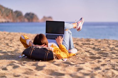 Millennial serbest çalışan kadın boş ekranlı ve deniz kenarındaki kumsalda yatan bir dizüstü bilgisayar kullanıyor. Rüya ofisi uzaktan çalışma konsepti 