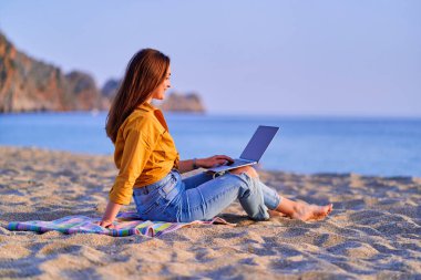 Genç mutlu, neşeli, tasasız, mutlu, binlerce yıllık özgür kadın bilgisayar kullanıyor ve deniz kenarında kumsalda oturuyor. Rüya ofisi uzaktan çalışma konsepti