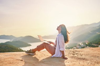 Ücretsiz mutlu serbest çalışan kız gün batımında güzel manzaralı bir bilgisayarın başında uzaktan çalışmaktan hoşlanıyor. Çevrimiçi her yerde çalışıyor 