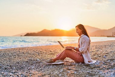 Happy smiling cute joyful satisfied girl freelancer remote working online on the seashore at sunset time. Work everywhere clipart