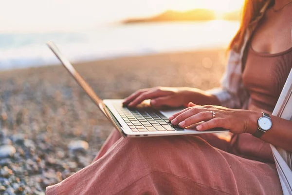 Close up of female freelancer typing on keyboard during remote working online on the seashore at sunset. Work everywhere