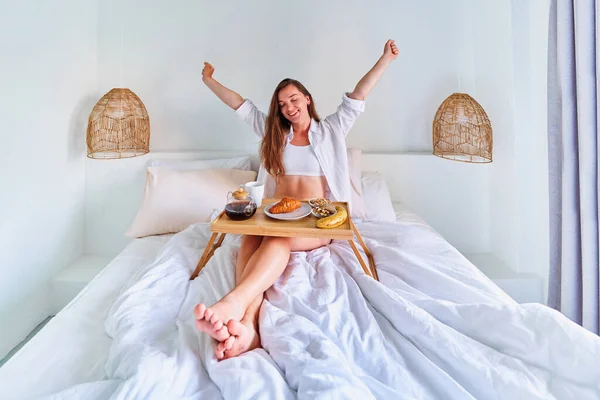 幸せな満足笑顔かわいい屈託のない喜び若い目覚めの女性は快適な怠惰な朝にベッドで朝食を楽しむ — ストック写真