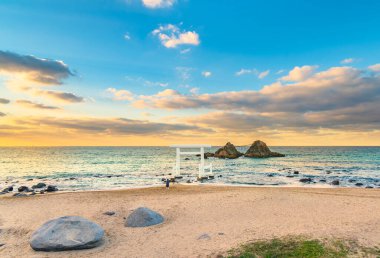 Fukuoka 'nın Itoshima plajında dua eden bir kişi beyaz bir Shinto torii kapısının önünde duruyor ve akşam günbatımında ünlü Sakurai Futamigaura' nın Meoto Iwa Çifti Rocks 'una çıkıyor..