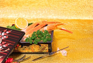 Japanese fresh raw king crab legs arranged in a luxurious box adorned with golden traditional motifs, and accompanied by seafood cutlery such as pliers or crab leg forks, against a gold background.