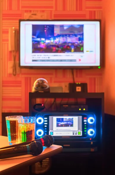 日本のカラオケクラブルームの2つのドリンクグラスの横にあるテーブルの隅に歌を歌うための2つの黒いマイクで 壁のテレビ画面の下のボタンにバックライト付きLedで点灯します — ストック写真