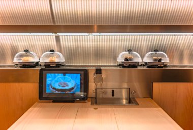 Japon Kaiten-zushi döner suşi restoranındaki ahşap bir masanın önü. Menü olarak kullanılan bir tablet ve şeffaf plastik bir kubbe tarafından korunan bir suşi tabağı taşıyan bir taşıyıcı bant..