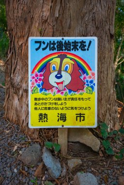 Shizuoka, atami - 26 Mart 2025: Bir ağaca eklenmiş Japon tabelasında renkli bir çizgi film köpeği ve hayvan sahiplerine kamu temizliği ve güvenliğini korumak için köpeklerinin pisliklerini temizlemelerini hatırlatan bir metin yer alıyor..