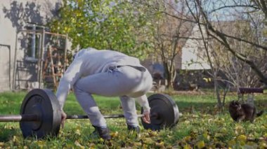 Siyah kedisi olan genç bir adam, kaslı vücudunu dışarıda eğitiyor. Sonbahar bahçesinde metal halterleri kaldıran vücut geliştirici. Yavaş çekim.