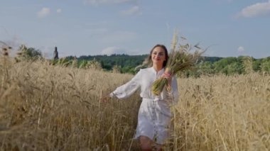 Beyaz elbiseli güzel bir Ukraynalı kadın elinde buğday başağıyla arka planda mavi bir gökyüzü ile buğday tarlasında koşuyor. Yavaş çekim.