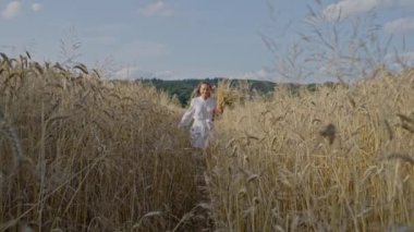 Beyaz elbiseli güzel bir kadın elinde buğday başağıyla arka planda mavi gökyüzü olan bir buğday tarlasında koşuyor. Yavaş çekim.