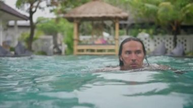  Uzun siyah saçlı yakışıklı beyaz adamın portresi havuzun yavaş çekimde video kameraya doğru yüzüyor. Yüksek kalite 4k görüntü