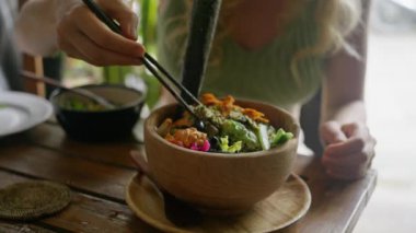 Çin yemek çubuklarıyla yemek yiyen bir kadın. Organik ve temiz yemek. Diyet öğle yemeğinde taze sebzeler. Avokado, fasulye, salatalık, zeytin, nori yosunlu salata, ahşap tabakta, sağlıklı yemek, ağır çekim