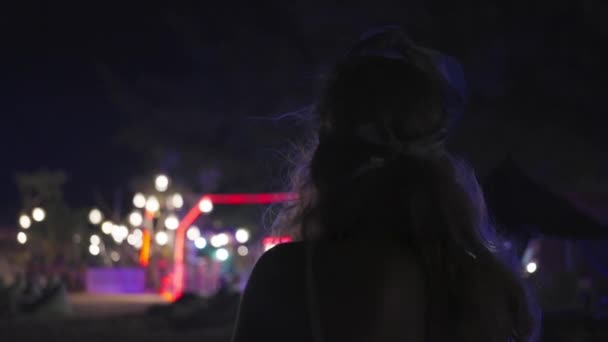 一个女人晚上站在海滩上 后视镜 孤独的概念 黄昏时分 在沙滩上的咖啡店 酒吧里的花环和灯笼的彩灯 平静的气氛 慢动作 — 图库视频影像