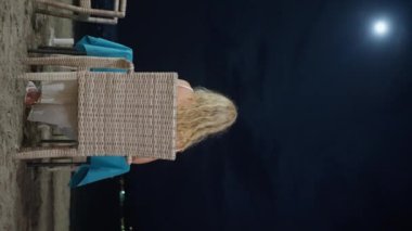Sarışın bir kız bir sandalyede oturuyor, okyanusun kıyısında, gece vakti. Geceleri okyanusun üzerinde dolunay. Ay ışığının güzel manzarası. Dikey video. Yüksek kalite 4k görüntü