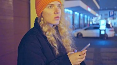 Turuncu Şapkalı Kadın. Ve siyah ceketli, caddede duran, bir kavşakta geceleyin, kışın, otelinin konumunu cep telefonundan izleyen. mobil uygulama kullan. 