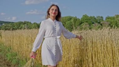 Beyaz elbiseli güzel Ukraynalı kadın elinde buğday başaklarıyla bir buğday tarlasında yürür arka planda mavi bir gökyüzü. Yavaş çekim.